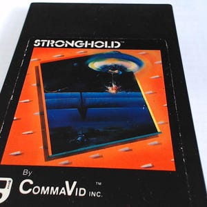 Puede incluir: Un cartucho de videojuego rectangular negro para Atari 2600 con el título STRONGHOLD en texto blanco. La etiqueta muestra un fondo naranja con una ilustración central de un paisaje azul, un platillo volante y el texto By COMMAVID INC.
