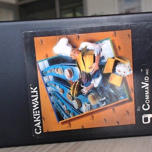 Könnte beinhalten: Eine schwarze, rechteckige Videospielkassette für das Atari Video Computer System mit dem Titel Cakewalk von CommaVid. Das Etikett zeigt eine Illustration eines Kochs mit weißer Mütze, der Kartons neben einem Fließband mit Kuchen und Gebäck hält.