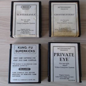 Könnte beinhalten: Vier rechteckige schwarze Kunststoff-Videospielkassetten für das Atari 2600-System. Die Etiketten zeigen die Titel Subterranea, Ghostbusters, Kung Fu Superkicks und Private Eye sowie Herstellerinformationen und Gebrauchsanweisungen.