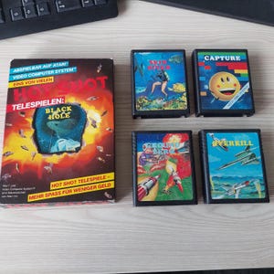 Pode incluir: Uma coleção de cartuchos de videogame vintage, incluindo um jogo Hot Shot em caixa intitulado Black Hole e quatro cartuchos soltos rotulados como Skin Diver, Capture, Ground Zero e Overkill. A caixa apresenta gráficos em vermelho e amarelo com texto em alemão.