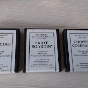 Könnte beinhalten: Drei schwarze, rechteckige Videospielkassetten für das Atari Video Computer System. Die Etiketten sind weiß mit schwarzem Text und zeigen die Titel Beamrider, Skate Boardin und Chopper Command sowie die jeweiligen Herausgeberinformationen.
