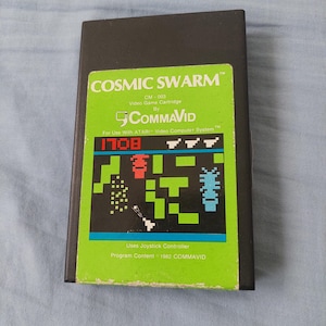 Può includere: Una cartuccia per videogioco Atari nera intitolata Cosmic Swarm di CommaVid. L'etichetta verde riporta il titolo del gioco, il testo CM-003 e una grafica pixelata che mostra una figura rossa, una creatura blu e una nave bianca che spara.