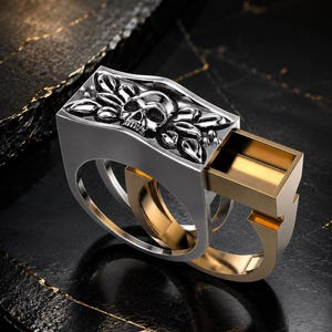 Puede incluir: Un anillo de dos piezas con una sección superior plateada que presenta un motivo esculpido de calavera y hojas. La pieza plateada se desliza sobre una banda interior dorada, revelando un compartimento rectangular hueco en la estructura metálica.