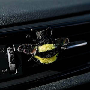 Op de afbeelding: Een pluizige bijvormige luchtverfrisserclip voor in de auto met zwarte en gele strepen, doorschijnende vleugels en dunne antennes, bevestigd aan de horizontale lamellen van een zwart ventilatierooster naast een rode alarmlichtknop.