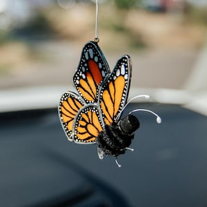 Puede incluir: Un adorno colgante que presenta una mariposa monarca de vidrieras con alas naranjas y negras, unida a un cuerpo negro difuso con dos antenas blancas delgadas. La mariposa está suspendida por un hilo blanco fino de un espejo retrovisor.