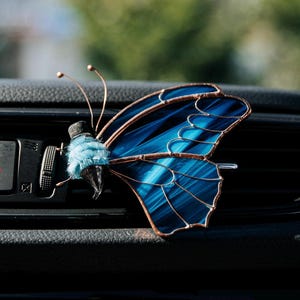 May include: A butterfly-shaped car air vent clip featuring stained glass wings in shades of deep blue and black with copper-colored metal soldering. The body is crafted from light blue fuzzy material with two thin copper wire antennae extending upward.