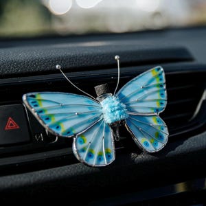 Peut inclure: Un désodorisant de voiture en forme de papillon avec des ailes bleu clair ornées de motifs circulaires jaunes et bleu foncé, bordées de métal argenté. Le centre présente un corps bleu clair duveteux et un petit flacon en verre avec un bouchon en liège.