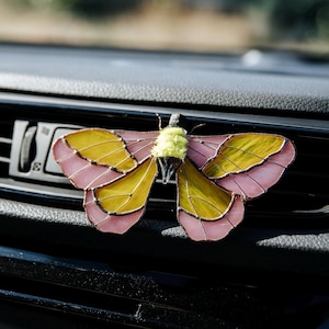 May include: A stained glass moth ornament featuring translucent yellow and pink wings with dark metal soldering lines. The center of the moth has a fuzzy, light green textured body and a dark, pointed tail section attached to a black clip.