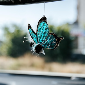 Puede incluir: Un adorno de mariposa de cristal tintado colgado de un espejo retrovisor. La mariposa presenta alas de color azul turquesa vibrante con bordes negros y puntos dorados, junto a un cuerpo negro difuso y antenas de alambre delicadas.