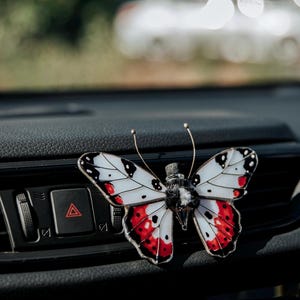Peut inclure: Un clip diffuseur de parfum pour voiture en forme de papillon en vitrail, avec des ailes blanches ornées de taches noires et de touches rouges sur les bords. Le corps central est en matière duveteuse noire et blanche avec deux antennes fines.