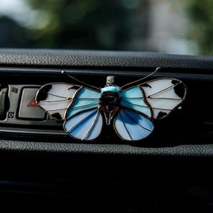 May include: A stained glass butterfly ornament featuring translucent white and light blue wings with dark edges and a fuzzy black centre body. The butterfly has thin wire antennae and is positioned against a dark textured dashboard surface.