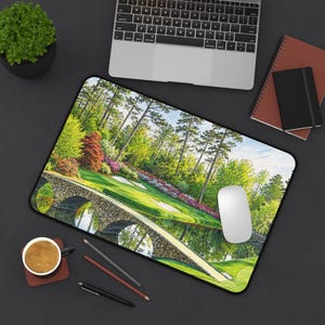 May include: A rectangular desk mat featuring a vibrant landscape print of a golf course with a stone bridge over a pond, lush green grass, tall pine trees, and colorful flowering bushes under a bright sky, with a white computer mouse on top.