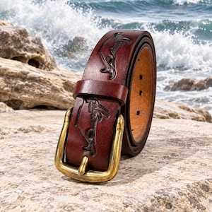 May include: A dark brown leather belt featuring embossed nautical imagery of a person fishing with a rod. The belt includes a brass-toned metal buckle and a matching leather keeper loop. The interior of the belt shows a lighter tan leather finish.