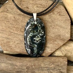 May include: Oval pendant necklace with a black and green design. The pendant is suspended from a black cord. The necklace is displayed on a wooden surface with driftwood accents. A white pot with a green plant is in the background.