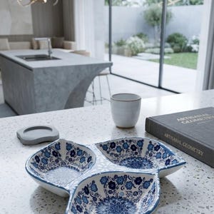 Può includere: Un piatto da portata in ceramica a tre sezioni con base bianca e complessi motivi floreali e mandala blu. Il piatto ha una forma curva a foglia con un bordo dipinto di blu, posizionato su un piano di lavoro bianco maculato.