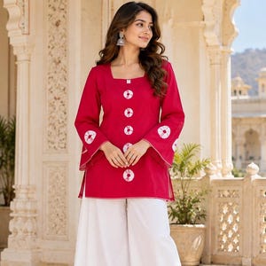 Puede incluir: Una túnica de color magenta vibrante con escote cuadrado, mangas largas con puños acampanados y una fila vertical de detalles circulares blancos de teñido anudado en el centro. Se combina con pantalones blancos anchos.