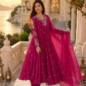 May include: A vibrant magenta Anarkali dress featuring long sleeves with cold-shoulder cutouts, intricate silver embroidery around the neckline and cuffs, and scattered small mirror embellishments throughout the flowing, pleated skirt fabric.