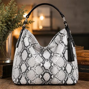 Pode incluir: Uma bolsa estilo hobo com um padrão de pele de cobra em branco e preto. A bolsa tem uma alça preta única fixada com ferragens prateadas e uma borla de couro preta pendurada na lateral. O material tem uma textura semelhante a escamas.