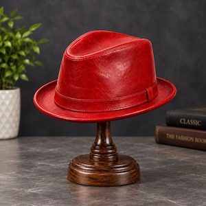 Pode incluir: Um chapéu fedora vermelho vibrante com um acabamento liso e polido e uma faixa vermelha a condizer à volta da copa. O chapéu é exibido num suporte de madeira castanho escuro, destacando a sua forma estruturada e aba curva.