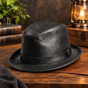 Könnte beinhalten: Ein schwarzer Fedora-Hut mit einer strukturierten, lederartigen Oberfläche. Der Hut verfügt über eine eingekniffene Krone und ein passendes schwarzes Band, das die Basis der Krone umschließt. Die Krempe ist leicht gebogen.