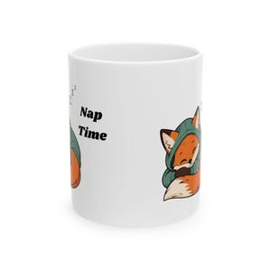 Könnte beinhalten: Ein weißer Keramikbecher mit einer Cartoon-Illustration eines schlafenden orangefarbenen Fuchses, der einen grünen Kapuzenpullover trägt. Der Text Nap Time ist in schwarzen Buchstaben neben dem Fuchs aufgedruckt, ergänzt durch zzz-Symbole.