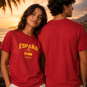 Può includere: Una t-shirt rossa a maniche corte con la parola ESPAÑA in lettere gialle sopra un grafico di una pallavolo, la bandiera spagnola e il testo LA FURIA ROJA con un piccolo simbolo di cuore giallo stampato sulla parte anteriore del petto.