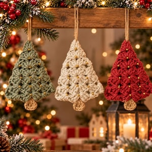 Könnte beinhalten: Drei gehäkelte Weihnachtsbaum-Anhänger in Salbeigrün, Creme und Rot. Jeder Baum hat ein strukturiertes Maschenmuster mit kleinen goldenen Perlen und einem kurzen braunen Stamm und ist an einer Schlaufe aus Juteschnur aufgehängt.