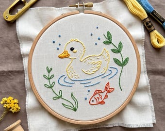 May include: A round wooden embroidery hoop containing white fabric with a hand-stitched design of a yellow duckling swimming in blue water, accompanied by a small orange fish, green aquatic plants, and blue bubbles, with gold scissors and thread.