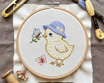 May include: An embroidery hoop containing a white fabric panel with a stitched design of a yellow chick wearing a blue hat with white flowers, accompanied by a blue butterfly on a green stem and a small pink butterfly, with embroidery thread and scissors.