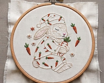 May include: A round wooden embroidery hoop containing white fabric with a hand-stitched design of a sleeping rabbit wearing a nightcap and pajamas patterned with small orange carrots. The rabbit holds a larger carrot, surrounded by tiny flowers.