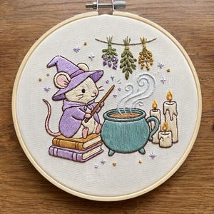 May include: An embroidery hoop containing a fabric panel with a stitched illustration of a mouse wearing a purple wizard hat and cape, sitting on two books, holding a wand over a teal cauldron with steam, three candles, and hanging herbs.