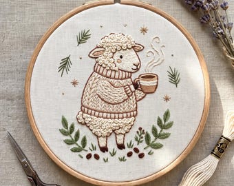 May include: An embroidery hoop containing a white fabric panel with a hand-stitched design of a sheep wearing a cable-knit sweater and holding a steaming mug. The scene includes green leaf sprigs, coffee beans, and small star accents.