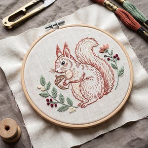 May include: A round wooden embroidery hoop containing a piece of off-white fabric with a hand-stitched design of a squirrel holding an acorn. The squirrel is rendered in shades of rust and cream, surrounded by green leafy branches with red berries.