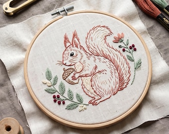 May include: A round wooden embroidery hoop containing a piece of off-white fabric with a hand-stitched design of a squirrel holding an acorn. The squirrel is rendered in shades of rust and cream, surrounded by green leafy branches with red berries.