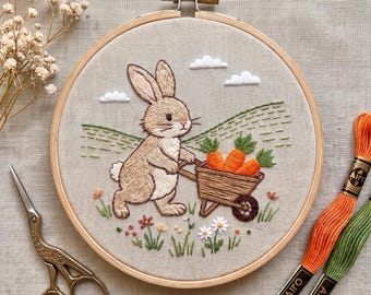 May include: An embroidery hoop containing a beige fabric panel with a hand-stitched design of a light brown rabbit pushing a wooden wheelbarrow filled with orange carrots. The scene includes green hills, white clouds, and small colorful flowers.