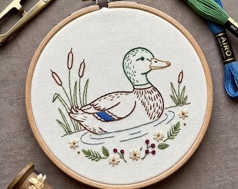 May include: A circular wooden embroidery hoop containing white fabric with a hand-stitched mallard duck design, surrounded by green reeds, small white flowers, and red berries. Includes gold metal embroidery scissors and two skeins of Airo thread.