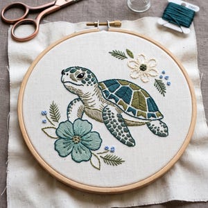 May include: An embroidery hoop containing a white fabric panel with a stitched sea turtle in shades of teal, olive, and cream. The turtle is surrounded by embroidered teal and white flowers and green leaves. Copper scissors, a thimble, and thread are nearby.
