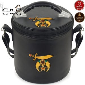 May include: A black cylindrical carrying case with a top handle and shoulder strap. The bag features embroidered gold-colored emblems of a scimitar sword over a crescent moon and star on both the top lid and the front side of the bag.