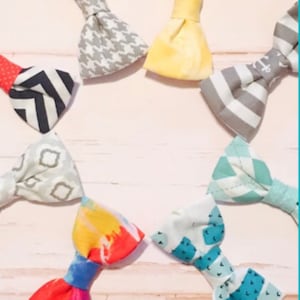 May include: Assortment of eight fabric bow ties in various patterns and colors, including gray, yellow, red, blue, and white. The bow ties are arranged in a circle on a light pink wooden surface.