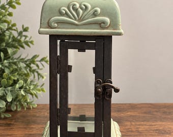 May include: A lantern featuring a light green ceramic top and base with embossed scrollwork and a dark brown metal frame. The central section has a hinged door secured by a small metal latch, and the ceramic surfaces show subtle brown shading.