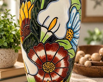May include: A tall, tapered ceramic vase featuring a glossy dark blue background adorned with a vibrant, hand-painted floral pattern. The design includes large yellow sunflowers, a white calla lily, a blue daisy, and a deep red flower with green leaves.