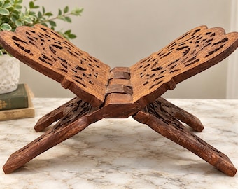 May include: A brown wooden book stand featuring an intricate, laser-cut floral pattern. The stand is constructed from two interlocking pieces that form an X-shape, designed to hold an open book with its carved panels angled upwards.