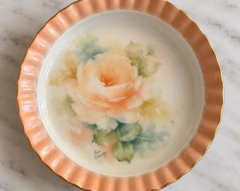 May include: A round porcelain dish with a fluted, peach-coloured rim and a gold-trimmed edge. The centre features a hand-painted illustration of a large, soft peach rose surrounded by muted green leaves against a white background.