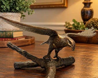May include: A bronze-toned metal sculpture of an eagle with wings spread wide, perched on a textured branch. The eagle is depicted in a downward-facing position with detailed feather carvings and a metallic finish, standing on a three-pronged base.