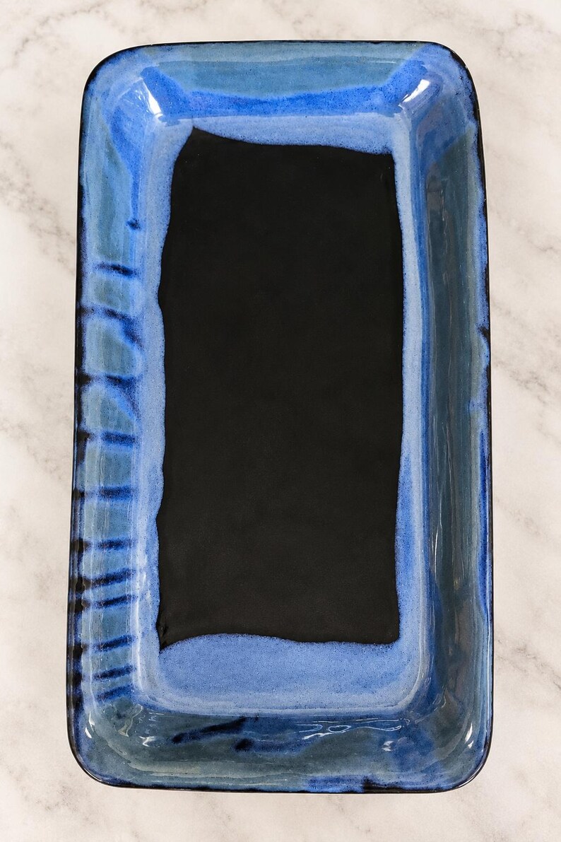 May include: A rectangular ceramic serving tray featuring a deep black center basin surrounded by a vibrant blue glazed border. The rim displays a reactive glaze effect with darker blue and black vertical brushstroke patterns along the edges.