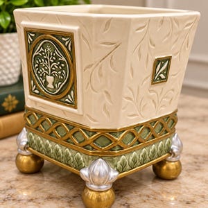 Puede incluir: Una maceta cuadrada de cerámica con cuerpo color hueso y patrones de hojas en relieve. Presenta un borde de celosía verde y dorado, un medallón central circular con un motivo de árbol y cuatro pies decorativos en tonos oro y plata.