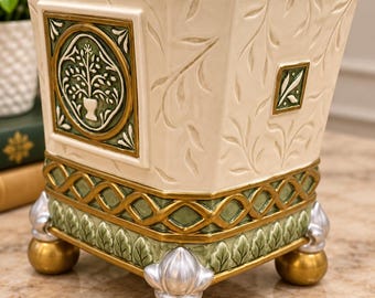 May include: A square ceramic planter with an off-white body featuring embossed leaf patterns. It has a green and gold lattice border, a central circular medallion with a tree motif, and four decorative feet finished in gold and silver tones.