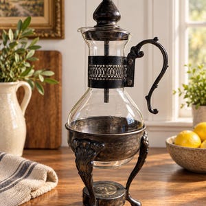 May include: A clear glass carafe with a dark, tarnished metal frame and base. The carafe features a decorative metal band around the neck, a curved handle, and a pointed metal lid. The base is supported by three ornate, curved metal legs.