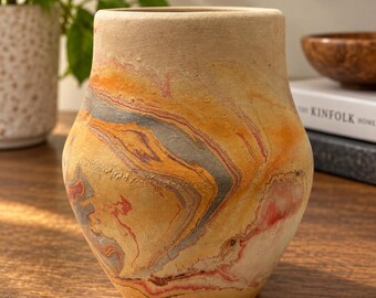 May include: A rounded ceramic vase with a matte, textured finish featuring a marbled pattern of swirling orange, yellow, red, and grey tones. The vessel has a wide, open top and a tapered base, showcasing a natural, earthy colour palette throughout.
