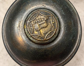 May include: A circular, dark-toned metal object with a weathered, oxidised finish. Centred on the top is a brass-coloured medallion featuring an embossed design of a stone wall structure and Hebrew text, enclosed within a decorative border.